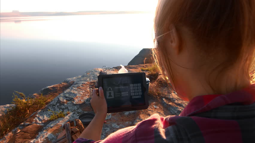 Young woman using tablet device for online internet banking, sitting next to tent at scenic beautiful lake on epic camping adventure.
