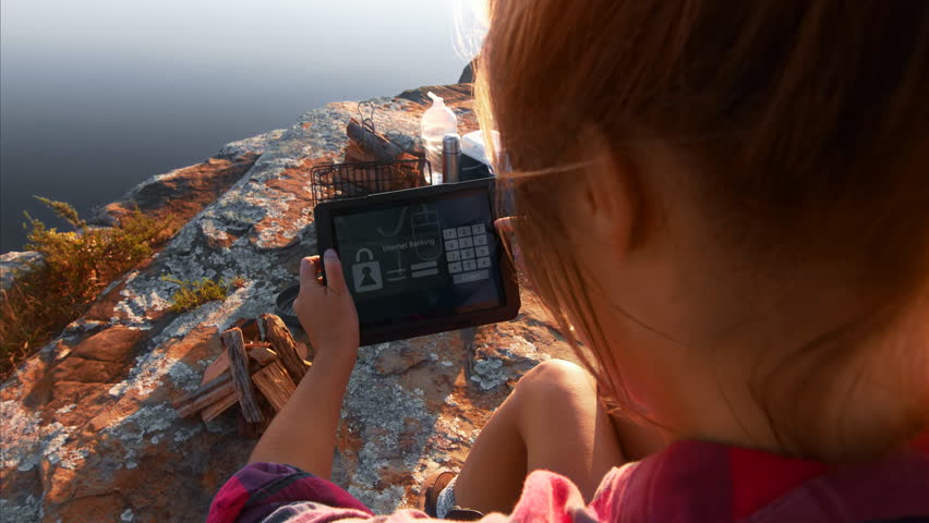 Young woman using tablet device for online internet banking, sitting next to tent at scenic beautiful lake on epic camping adventure.
