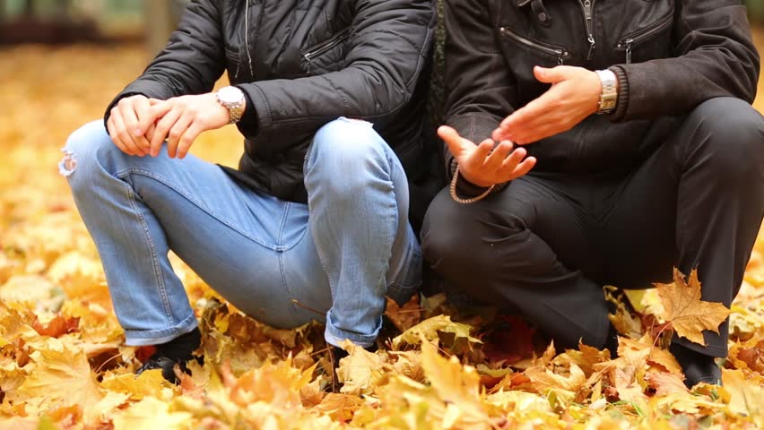 Two men sit on their haunches beside a tree in autumn park, stand up and leave