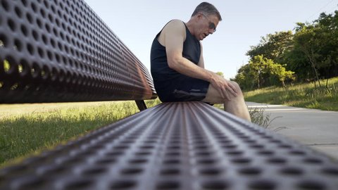 Middle aged Caucasian man with knee pain rests on a jogging path bench during outdoor exercise effort 4k