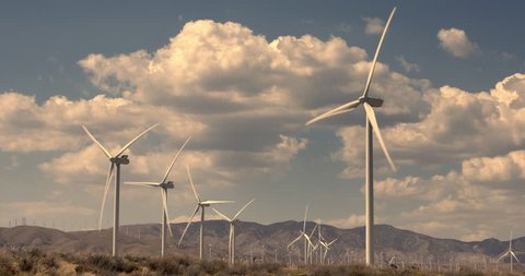 Wide view of large wind turbines tinted with golden light in Mojave Desert at Tehachapi in California.  Lots of puffy cumulus clouds and mountains visible in background.