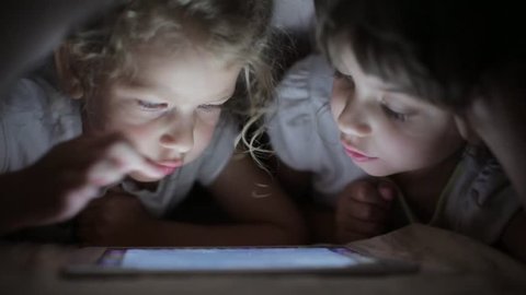 two little girls sisters draw on a tablet PC hiding under a blanket