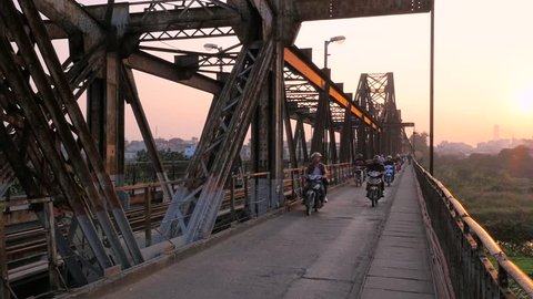 Motorbikes during rush hour in the evening sunset at the old Long Bien bridge in Hanoi. Motorbikes, pedestrians and trains cross this colonial french heavy bombed iron structure. Wide shot.