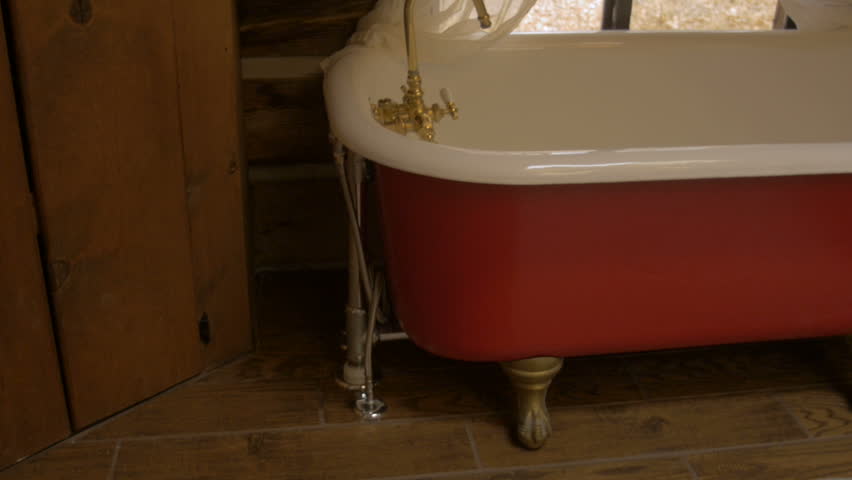 Red and white claw foot tub in a log cabin with white curtains and a window looking outside to dead leaves - pan up dolly shot