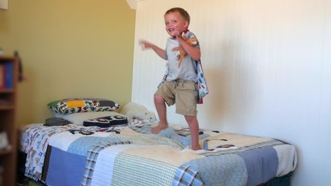 A slow motion shot of a cute happy little boy jumps on his bed while playing in his bedroom with his super hero cape