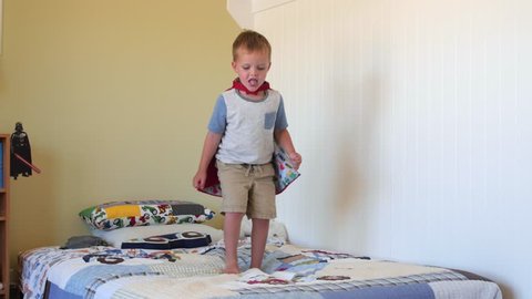 A cute happy little boy jumps on his bed while playing in his bedroom with his super hero cape