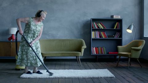 happy elderly woman listening music on headphones and dancing with a vacuum cleaner, home fun