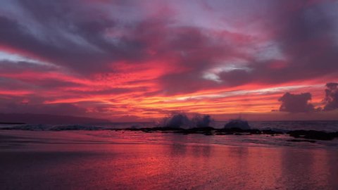 SLOW MOTION: Enormous powerful and wide ocean wave crushing into rocky reef. Water drops splashing and falling onto smooth sandy beach under stunning pink and purple summer evening skies