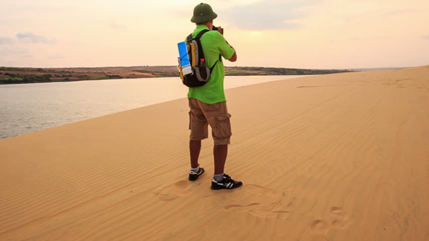 closeup european man in green hat with small backpack stands on dune by lake takes photos boundless white sand dunes at sunset in Vietnam