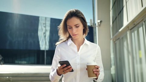 Beautiful young businesswoman wearing white shirt and using modern smart phone while walking at break in the city, professional female employer typing text message on cellphone outside, slow motion
