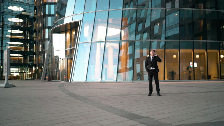 Funny joke. Young businessman have a smartphone talking. Goof stupid competitor running and giving kick ass to suit clothes man and runs away. Businessman overtake rival. Glossy business centre | Shutterstock HD Video #17526178