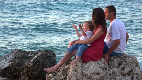 Young parents and happy child sitting together, rocky seashore splashing waves