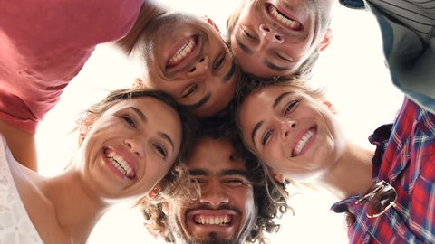 Directly below shot of young friends forming huddle. Low angle view of girls and guys with their head forming a circle. Portrait of young people looking at camera. Friendship and unity concept.