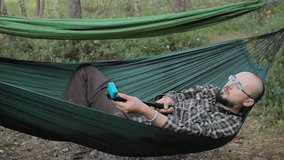 A man in a hammock making video on selfie stick with action camera. Man in the woods with a beard and glasses