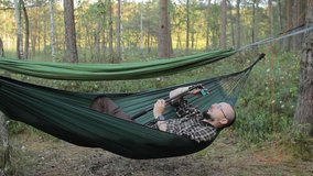 A man in a hammock making video on selfie stick with action camera. Man in the woods with a beard and glasses