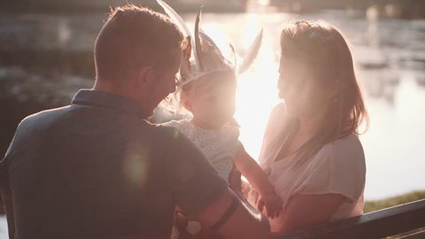 Young family with Baby Girl Enjoying Sunny evening in the Park. Slow Motion 120 fps, 4K. Parents Kissing Toddler Daughter in both cheeks, sitting on the near lake. Happy childhood and Parenthood.