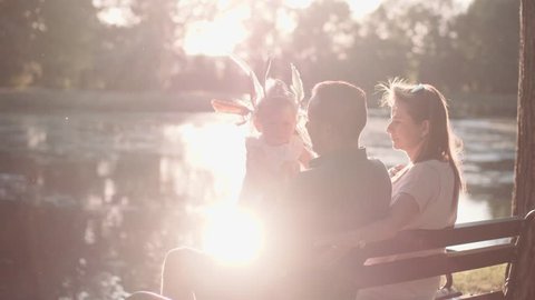 Young family with Baby Girl Enjoying Sunny evening in the Park. Slow Motion 120 fps, 4K. Parents playing with Toddler Daughter, sitting on the bench near lake. Happy childhood and Parenthood. Love