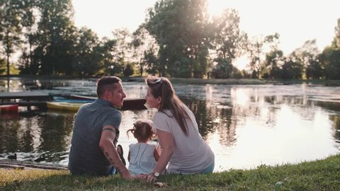 Young family with Baby Girl Enjoying Sunny evening in the Park. Slow Motion 120 fps, 4K. Parents kissing, while sitting with their toddler daughter near lake. Happy childhood and Parenthood. Love