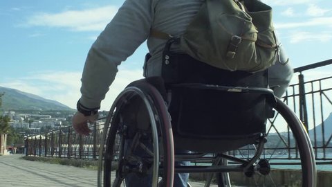 Slow motion shot of a traveling man with a backpack in wheelchair walking on the embankment along the sea