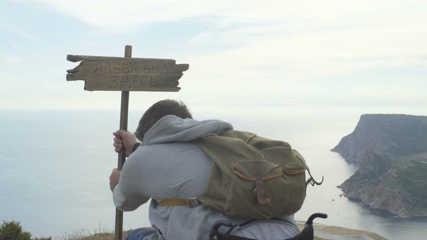 Young man in wheelchair on the top of mountain sets the plate with the inscription Alba was here and look at the sea