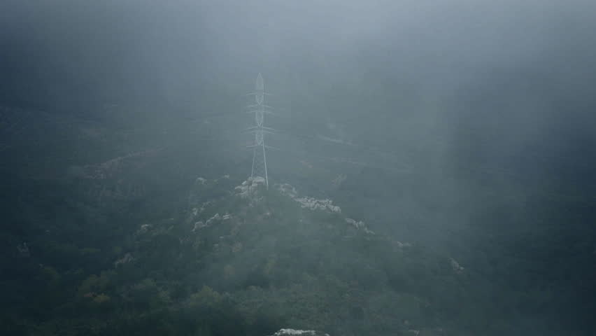 Birdsight from top of the mountains down on forest below and power transmission line support, strong wind blows on clouds that go very low and make thick fog