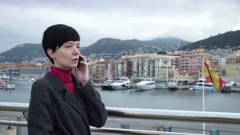 Female in winter season standing in harbour with yachts and boats. Elegant caucasian model wearing in coat using app on smartphone in mediterranean country. On the background sea view city and port