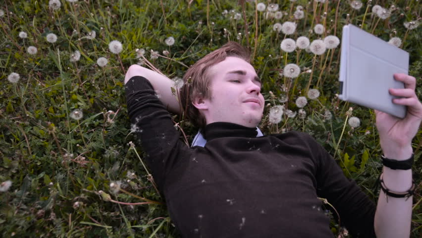 Young happy student using tablet in the park, lying on the grass in dandelions, smiling and laughing.