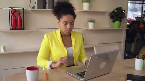 African businesswoman working on the computer in modern room with creative interior. Female concentrated on her job. Multicultural model young and professional wearing in formal yellow jacket. 