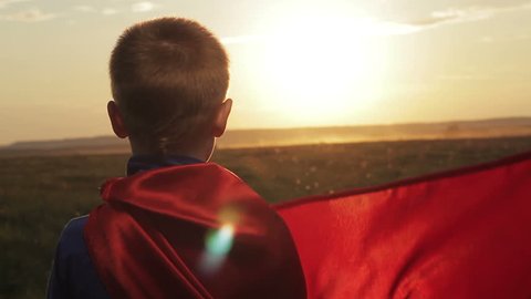Boy dressed with a Superman cape running in a field, looking into the sunset. Superhero kid flying on sunset sky background. Power concept. Child pretending to be superhero.