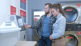Young cheerful couple doing laundry together at laundromat shop in 4k UHD video.
