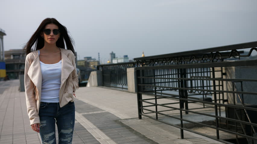 Young serious brunette woman in white jacket, blue jeans and black sunglasses walking on the modern bridge.
