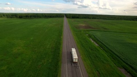 Aerial view camera flies following white cargo truck moving patched bad countryside road between green field - slow and high video footage shot with drone quad copter 4K HD