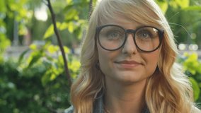 Closeup portrait of young beautiful blonde woman wearing glasses at summer green park. Closeup. RAW video record.