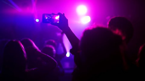 A person recording video with his phone in Club. Show with crazy lights. Crowd making party at a rock concert. Hands hold phone among people at rock concert