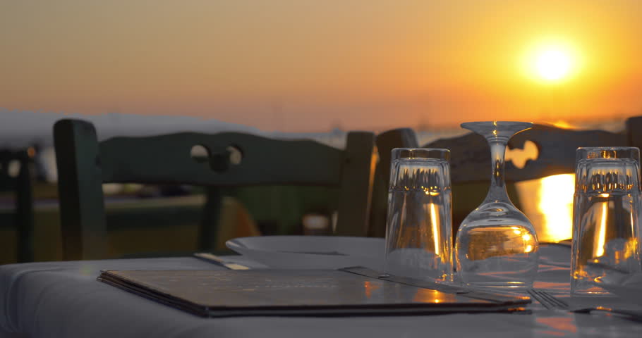 Empty table in outdoor restaurant at sunset. Served dishware and glassware with menu on white tablecloth. Sea and golden sundown in background