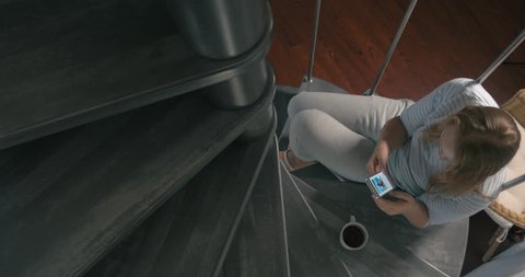 Cinemagraph - high-angle shot of young woman sitting on the winding stairs and watching newsfeed in her smartphone Video stock