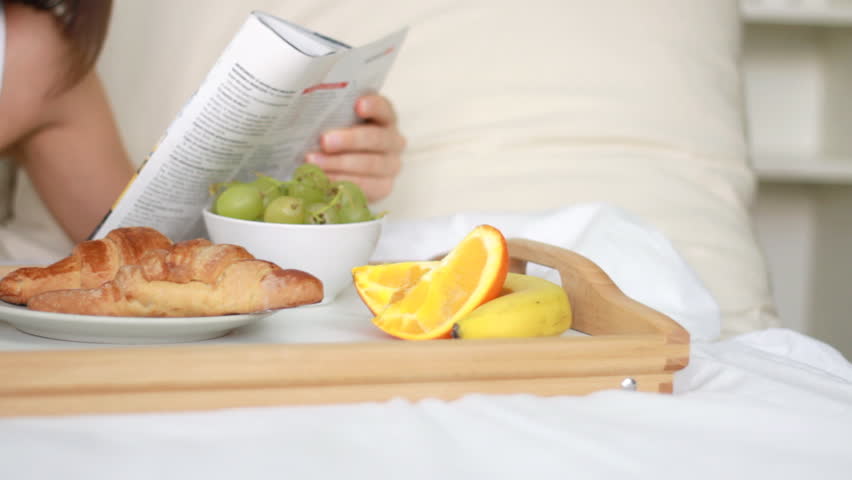 Woman eating breakfast in bed and reading magazine, dolly shot