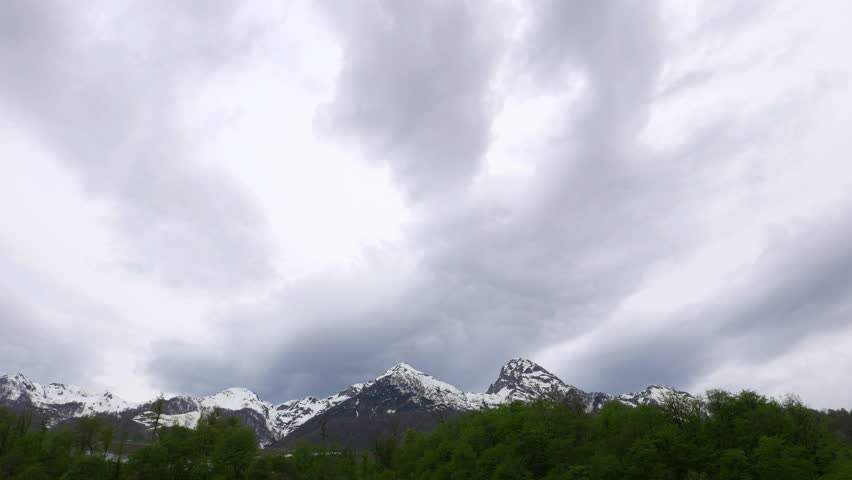 Gray clouds fly towards over rocky mounts, time lapse shot, low angle view. Rainy overcast, continuous clouds over sky. Light snow cover sharp peaks at top, green forest seen lower