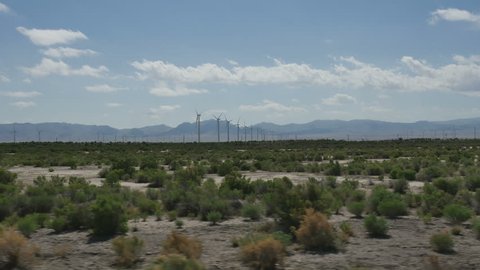 GREAT BASIN DESERT, WESTERN UTAH-JUNE 2016: POV-Driving side window view-Passing rows of electrical generating wind turbines turning with clouds in the sky and far distant mountains in desert scene.