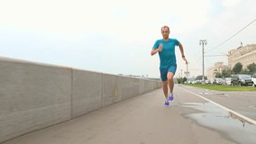 Sportsman in blue uniform running at camera on river embankment after rain against parapet and cloudy sky. 4K steadicam video