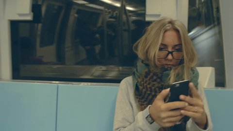  Young female hipster woman sitting in subway in Hong Kong, using smart phone. Pretty blonde teenage girl  with long hair, wearing  glasses and huge scarf going in underground.