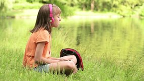 Little girl with tablet pc and headphones listening to music or watching video in summer park at lakeside.  Child have rest near water in park.