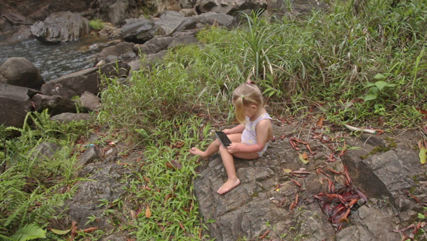 little blond girl with pigtails in swimsuit sits on stony river bank plays with phone against waterfall