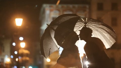 Romantic noir-styled couple under rain kissing on night city street, hiding from rain under umbrellas. City lights show pair silhouettes