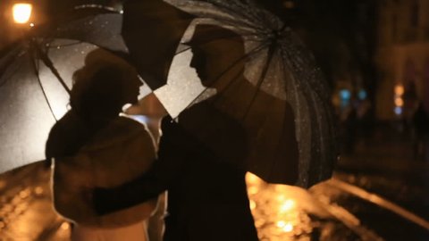 Beautiful couple walks under rain on night city street, hiding from rain under umbrellas. City lights show pair silhouettes
