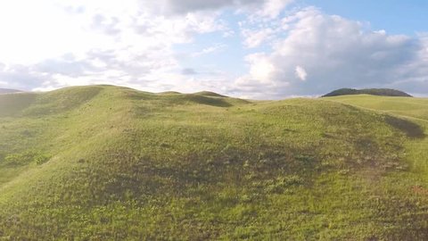 Drone aerial footage of flying above a rolling grassland hills on a sunny day with some beautiful clouds. The camera flies above the relaxing prairie like setting, with only nature to be seen.