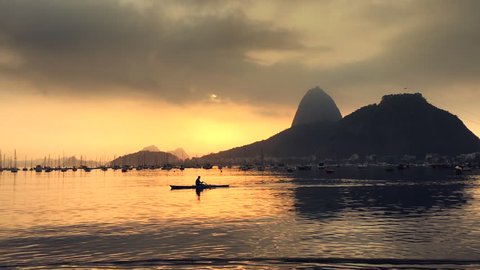 A lone rower passes in front of a golden sunrise silhouette of Sugarloaf Mountain at Botafogo Bay in Rio de Janeiro, Brazil