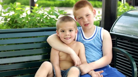 Brothers sit together on a park bench, the older one holding his younger brother.