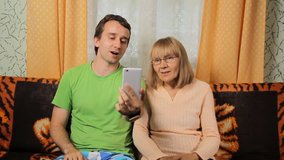 Adult son and elderly mother making video call via telephone. Mother and son smiling at home on sofa
