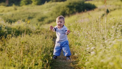 Baby makes first steps hesitantly. In Nature, standing on a path among the greenery and wildflowers. On the Sunset.